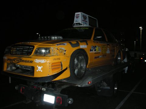 1992 Ford Crown Victoria - A heavily modified yellow sedan with various decals is being transported on a flatbed truck at night.