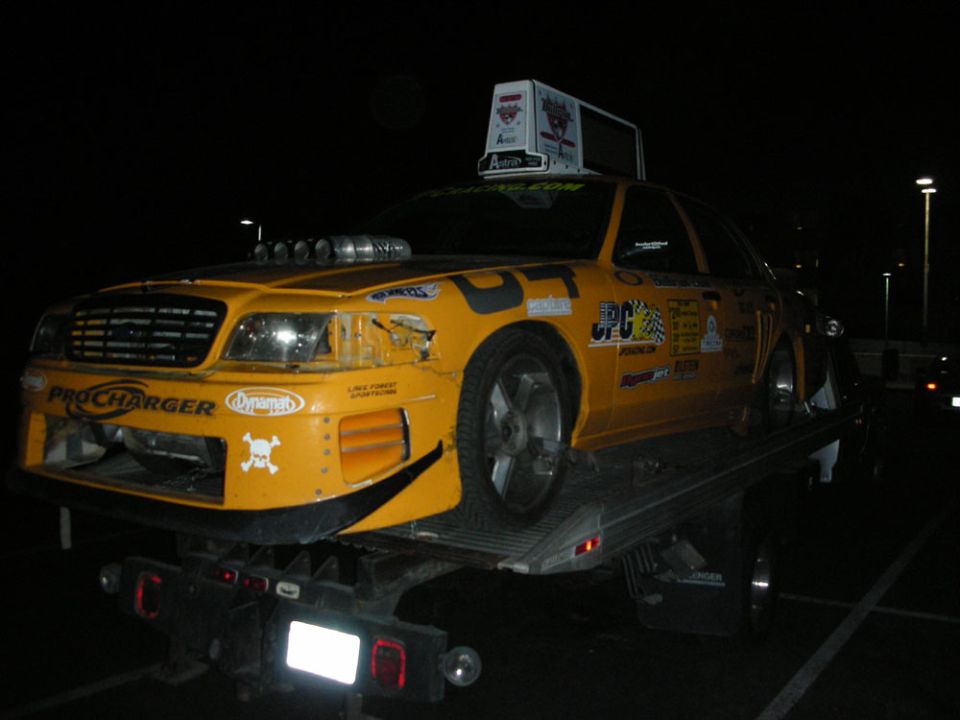 Damaged yellow race-liveried Ford Crown Victoria loaded on a flatbed tow truck at night