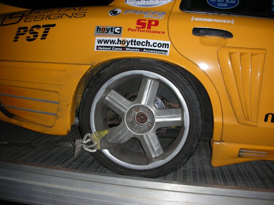 Yellow car with racing stickers, custom wheels, and tie-down straps on a trailer