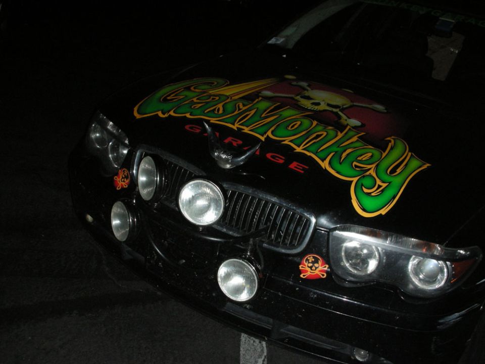 Black car front with Gas Monkey Garage skull and crossbones decal on hood, extra fog lights mounted on grille