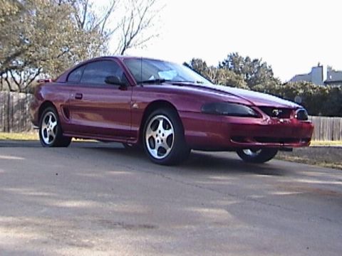 1989 Ford Mustang - This red coupe is parked on an asphalt surface, showing its side profile.