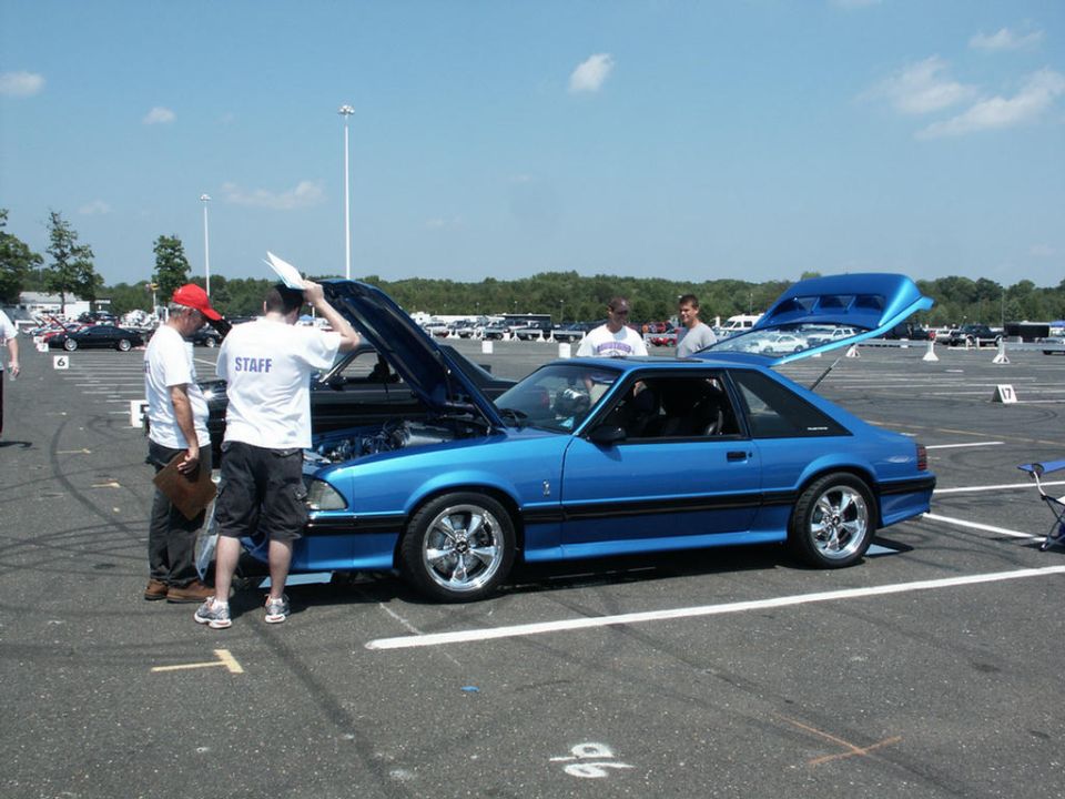 Blue coupe with hood and trunk open, people looking at engine. Looks like a Ford Mustang