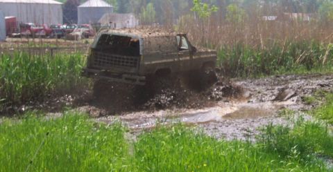 1978 Ford Bronco - This SUV is navigating a challenging muddy terrain, with tall grass and farm buildings in the background.
