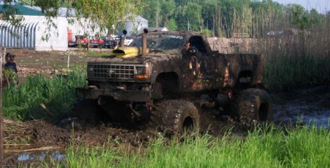 1978 Ford Bronco - A heavily lifted Ford pickup, completely coated in wet brown mud, climbs out of a bog pit on huge cut agricultural tires with a yellow-topp…