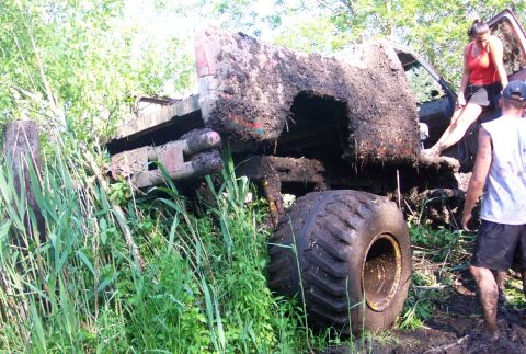 1978 Ford Bronco - A mud-plastered lifted 4x4 rests tipped on its side in tall grass beside a mud hole, its exposed underside showing frame rails, driveshaft…