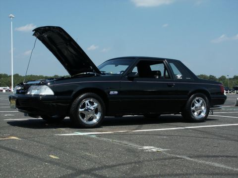 2001 Mercury Grand Marquis - A black coupe, likely a Ford Mustang from the early 90s, is parked with its hood open under a clear sky.