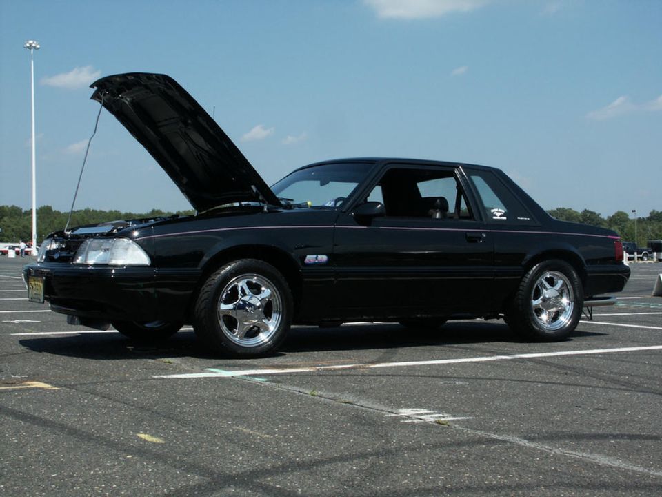 black coupe, hood up, parked in lot. looks like a fox body Mustang