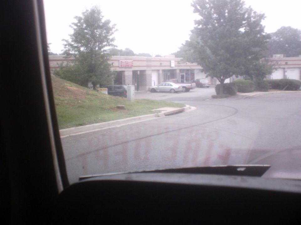 View through windshield over dark hood toward parking lot and commercial building; reversed 'FIRE DEPT' text on glass