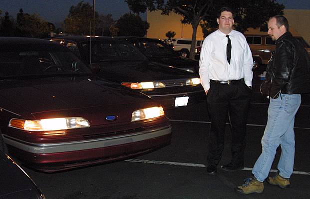 Maroon Ford sedan with headlights on in a parking lot at dusk, two men standing nearby