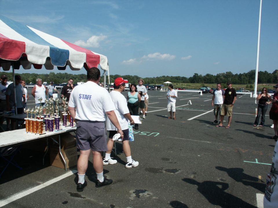 Car show event with trophies on a table under a tent, staff members present, and vehicles parked in the background
