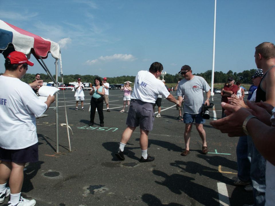 People gathered under a tent and on a paved lot, with a white pole in the background