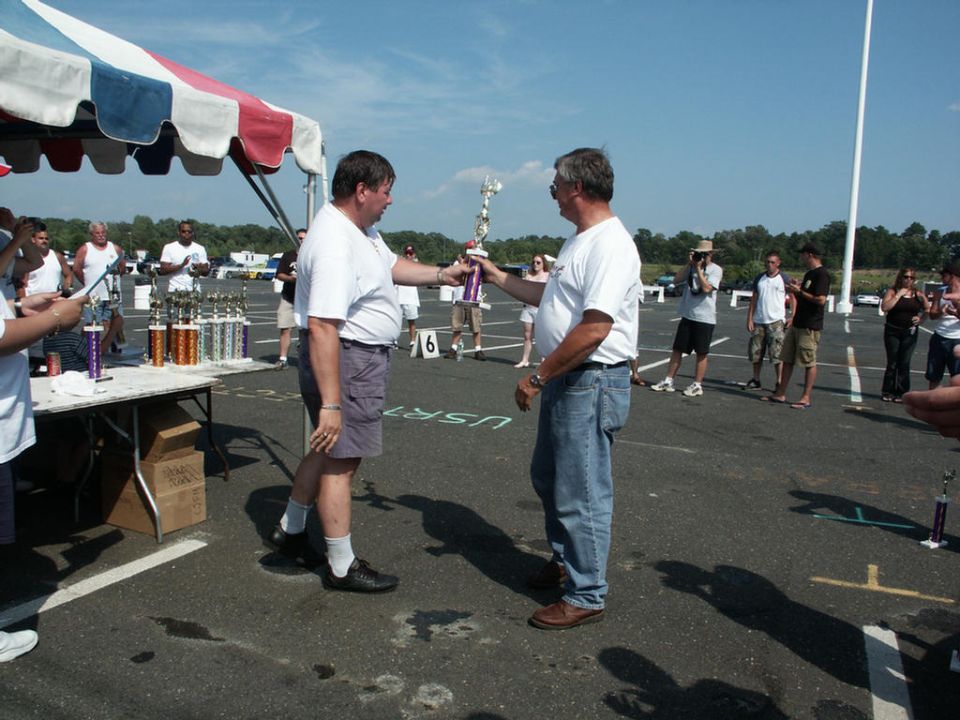 Two men exchange a trophy at an outdoor car show awards ceremony