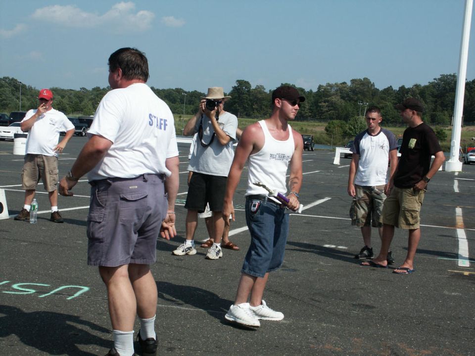 Group of men standing around in a parking lot, one taking a photo. A 'Staff' shirt is visible