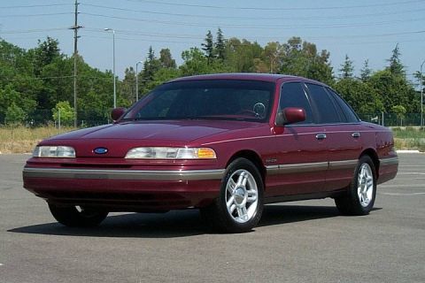 1992 Ford Crown Victoria - A maroon/dark red 1990s Ford Crown Victoria full-size sedan, shown from the front three-quarter angle, parked in an empty paved lot with tr…