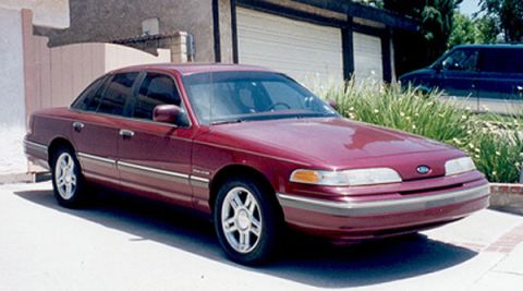 1992 Ford Crown Victoria - A dark red/maroon four-door full-size sedan photographed from the front three-quarter angle, parked on a driveway in front of a garage with…