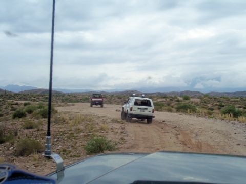 1991 Ford Bronco - Two SUVs, one white and one red, travel on a dirt road through a dry, hilly landscape.