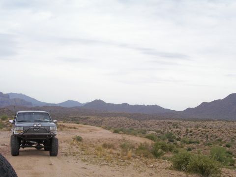 1991 Ford Bronco - A modified dark SUV is parked on a dirt road, with a mountainous desert landscape behind it.