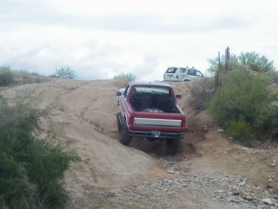 Red pickup truck with lifted suspension driving on a dirt trail, white SUV in background