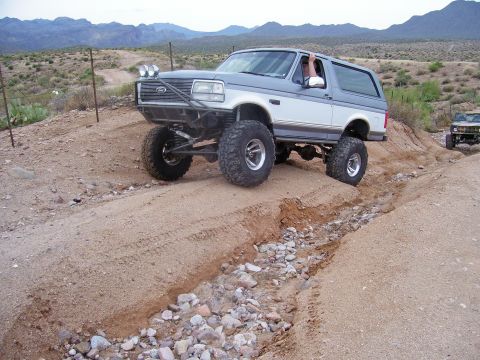 1991 Ford Bronco - A lifted 1991 Ford Bronco navigates a rocky dirt path in a desert landscape, with another off-road vehicle following.