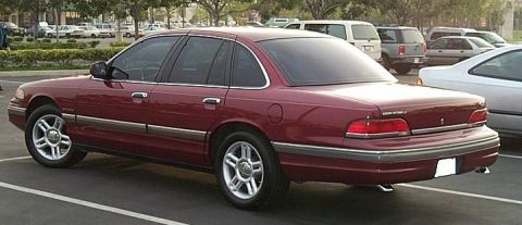 1992 Ford Crown Victoria - A dark red/maroon Ford Crown Victoria four-door sedan viewed from the rear left three-quarter angle, showing the 'CROWN VICTORIA' badge on…