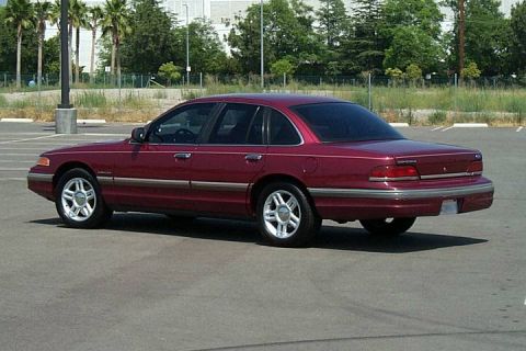 1992 Ford Crown Victoria - A dark red/maroon four-door full-size sedan photographed from the rear three-quarter left angle in an empty asphalt parking lot, with alloy…