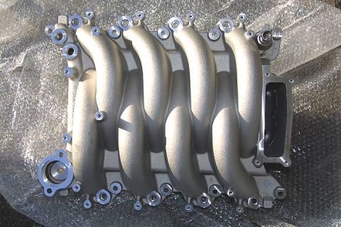 1992 Ford Crown Victoria - A cast-aluminum V8 intake manifold with long curved runners and multiple bolt bosses, photographed top-down on bubble wrap. The bare silver…