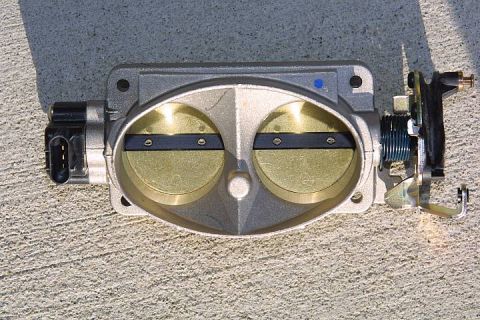 1992 Ford Crown Victoria - A twin-bore aluminum throttle body photographed face-on against concrete, showing both gold-anodized butterfly plates closed on the shaft.…