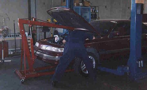 1992 Ford Crown Victoria - A dark red full-size sedan parked on a two-post shop lift with its hood open while a person in blue coveralls leans into the engine bay. A…