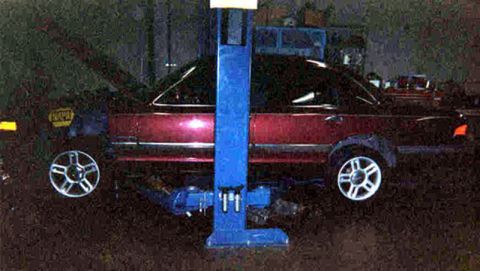 1992 Ford Crown Victoria - Dark burgundy Ford sedan raised on a blue two-post lift in a dimly lit shop, with two five-spoke alloy wheels sitting on the floor beneath…