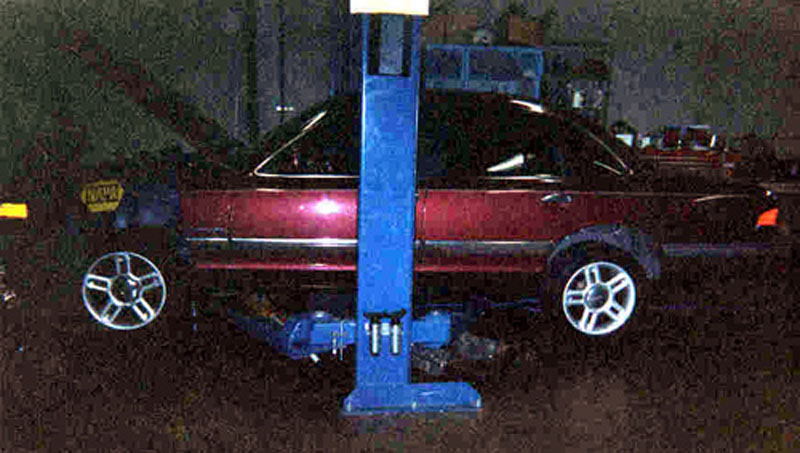 Burgundy sedan raised on a blue two-post lift in a dark shop with alloy wheels on the floor below