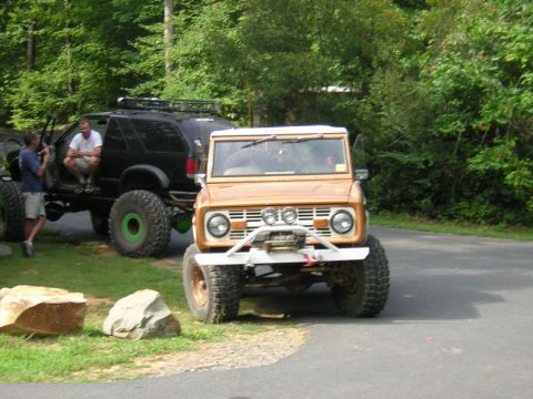 1979 Ford Bronco - A lifted orange Ford Bronco is parked on a paved surface next to a black SUV with large tires.