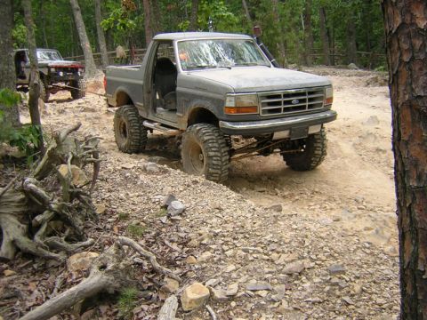 1979 Ford Bronco - Two modified trucks navigate a challenging off-road trail through a wooded area.