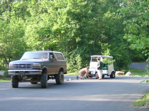 1979 Ford Bronco - A lifted Ford Bronco is parked on a road next to a golf cart with people working on it.