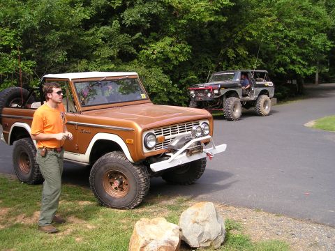 1996 Ford Bronco - A pair of classic Ford Broncos, one bronze and one black, are parked on a paved surface with a wooded background.