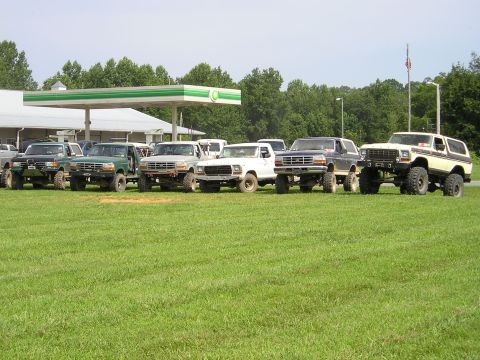1996 Ford Bronco - Several lifted Ford trucks and Broncos are parked in a grassy area near a gas station.