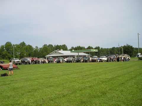 1996 Ford Bronco - Several SUVs and trucks are gathered in a field with a gas station in the background.