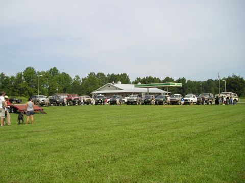 1996 Ford Bronco - A collection of SUVs and trucks are gathered in a field on a sunny day, with a gas station visible in the background.