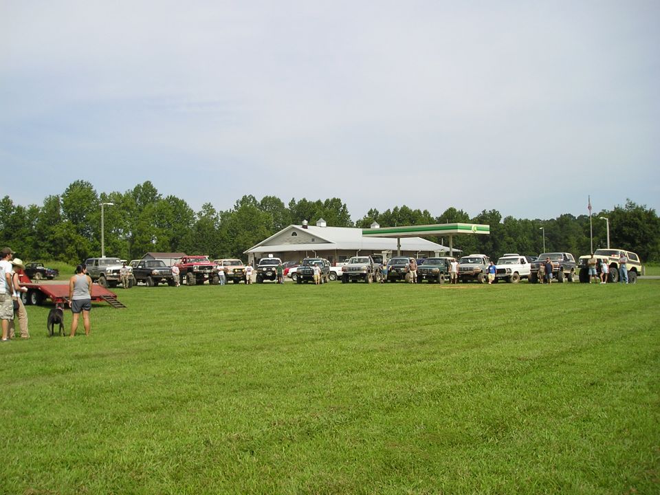 Group of SUVs and trucks parked in a grassy field near a gas station