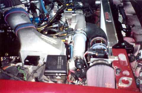 1992 Ford Crown Victoria - Engine bay of a red car fitted with a centrifugal supercharger: the polished blower head and its drive pulley feed a large intake tube into…