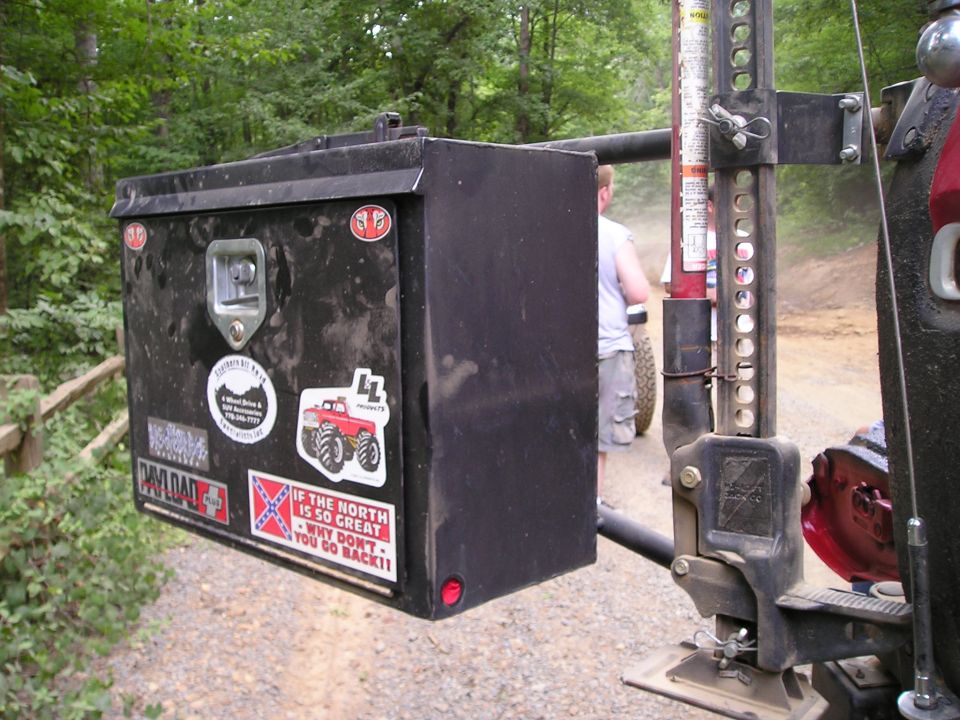 Black toolbox with stickers, attached to a vehicle with a Hi-Lift jack. Another person and vehicle are visible in the background