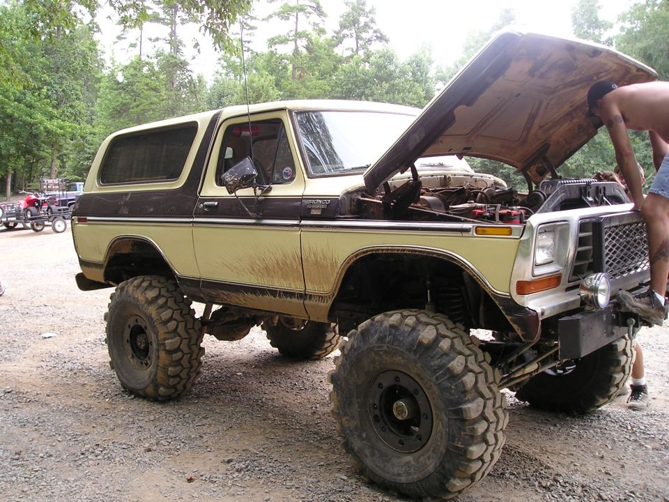 Lifted Ford Bronco with hood open, person looking at engine. Other vehicles and trailers in background