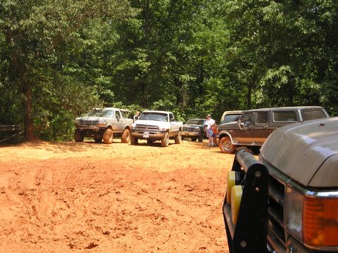 1996 Ford Bronco - Several off-road vehicles are gathered in a dirt lot surrounded by trees.