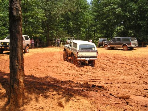 1996 Ford Bronco - Several off-road vehicles navigate a muddy terrain under a canopy of trees.