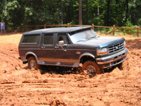 1996 Ford Bronco - A dark blue 1996 Ford Bronco navigates through deep mud, with its front-right wheel partially submerged.