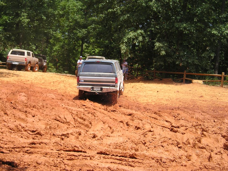 Rear view of a dark gray Ford Bronco driving through mud, with another tan Bronco and a dark SUV in the background