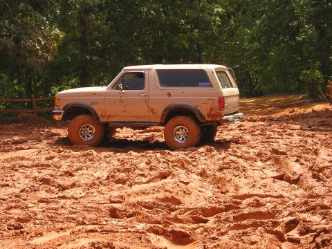 1996 Ford Bronco - A tan Ford Bronco navigates a challenging muddy terrain with dense foliage visible behind it.