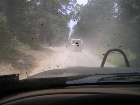 1996 Ford Bronco - Two SUVs travel on a dusty dirt road surrounded by trees.