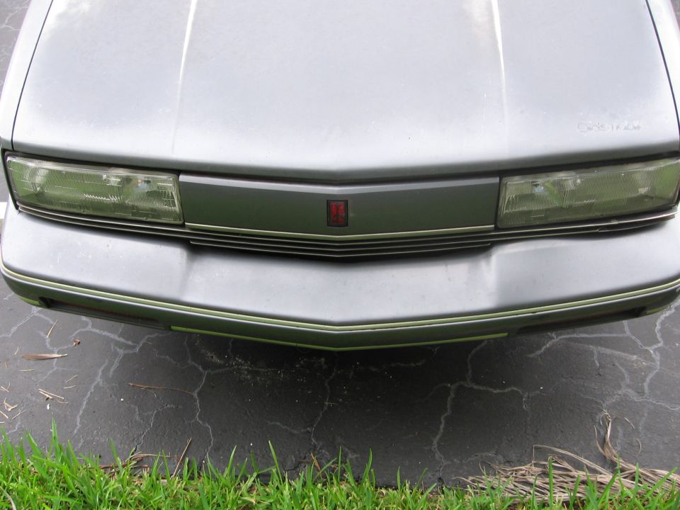 Front end close-up of a silver Oldsmobile Calais showing grille, headlights and red Olds emblem