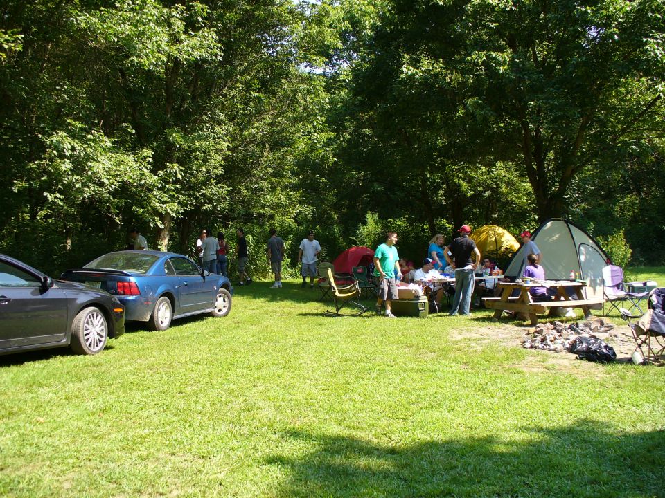 Blue coupe parked on grass, side view. People and tents in background