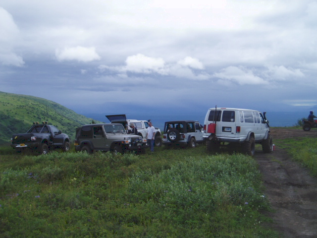 White van with red gas can on rear, parked on a dirt road with other off-road vehicles